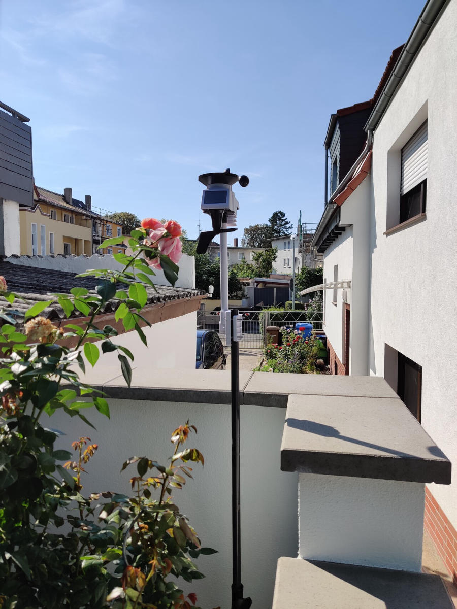 Ein sonniger Tag über einer ruhigen Nachbarschaft, mit einem Wetterstation auf einem Ständer, der auf einem Balkon steht. Im Vordergrund blühen rosa Rosen, während im Hintergrund ein Blick auf mehrere Häuser und Gärten zu sehen ist.