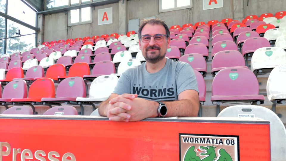 Christian Mpnch sitzt lächelnd auf einem Platz in einem Stadion, mit einem T-Shirt, das den Schriftzug „WORMS“ trägt. Im Hintergrund sind leere, farbige Sitze der Tribüne zu sehen, die in verschiedenen Farben angeordnet sind, und ein Schild mit der Aufschrift „WORMATIA 08“ ist ebenfalls sichtbar.